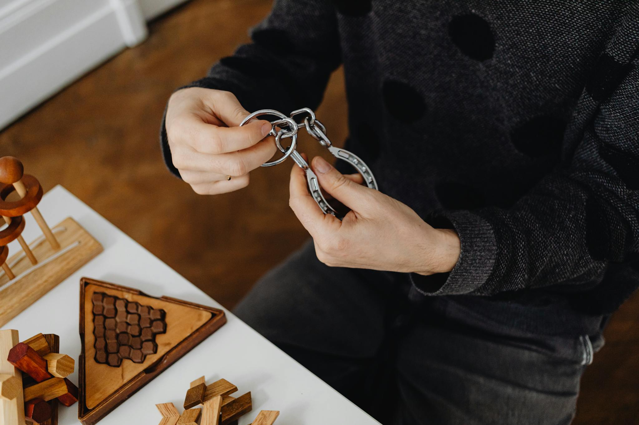 Top view of hands solving metal puzzle, surrounded by wooden brain teasers on a table.